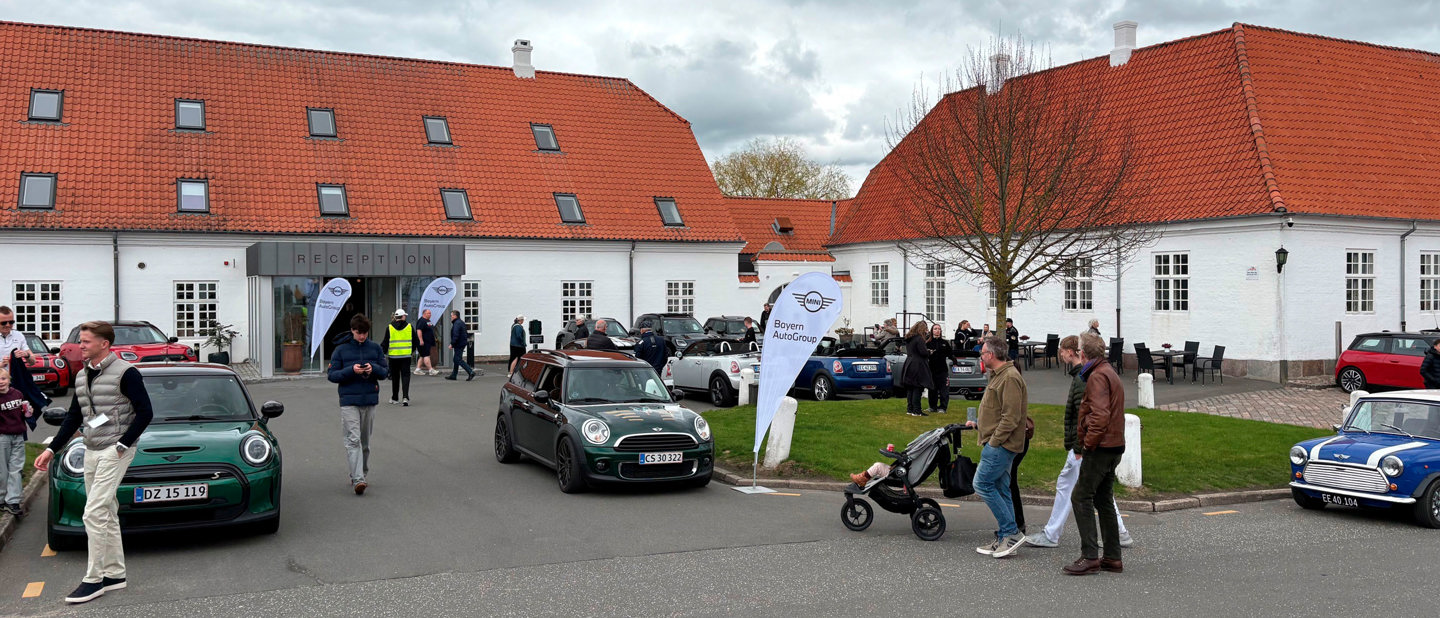 Folk samles uden for en hvid bygning til MINI Spring drive omgivet af parkerede Mini Coopers og to BAG-flag.