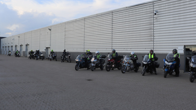 En række mennesker med hjelme og sikkerhedsveste sidder på motorcykler, der er parkeret ved siden af en stor industribygning.