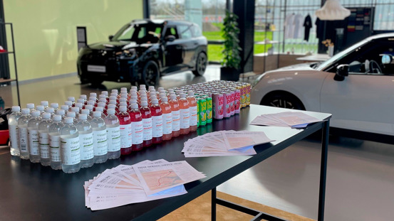 Et bord med sodavand og brochure  er arrangeret i et showroom med to biler udstillet i baggrunden.