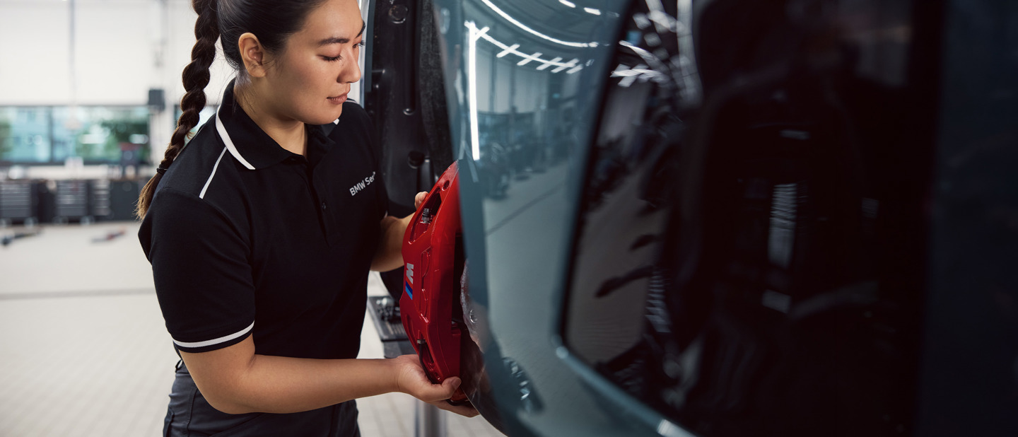 En BMW mekaniker i sort poloshirt inspicerer eller reparerer en bils røde bremsekaliber i et rent, veloplyst værksted.