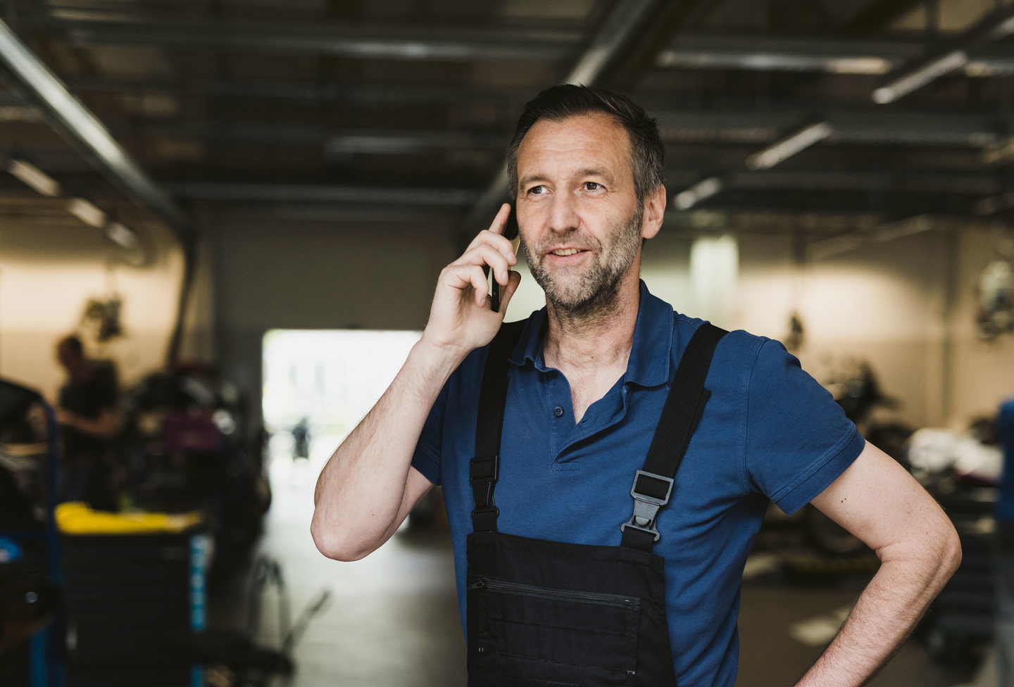 En midaldrende mand i arbejdsdragt taler i en smartphone på et værksted eller i industrielle omgivelser.