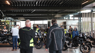 Folk i motorcykeltøj står og snakker mellem motorcyklerne i et showroom med metalloft og store vinduer.