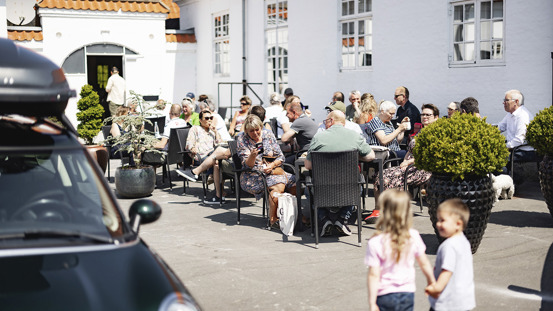 Mennesker siddende på caféstole ved en hvid bygning. 