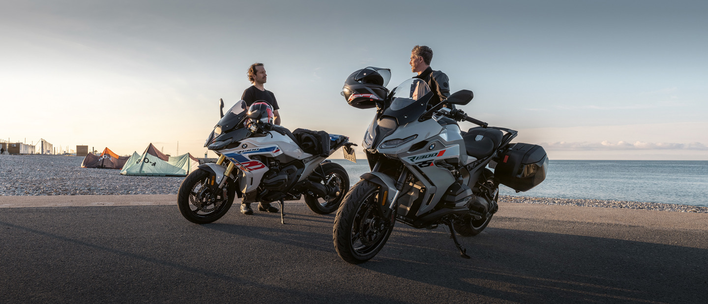 To personer står ved parkerede motorcykler på en vej nær stranden, med telte og havet i baggrunden.