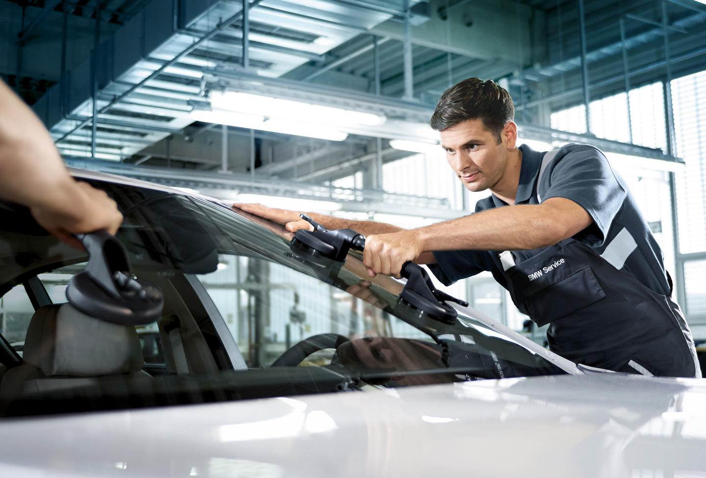 En mekaniker i BMW-uniform bruger sugeværktøj til at montere eller justere en forrude på et veloplyst autoværksted.