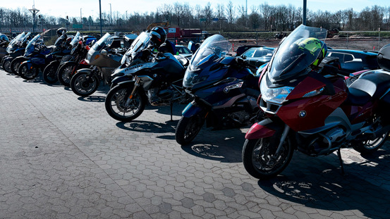 En række parkerede BMW Motorrad motorcykler står på et asfalteret område med træer og biler i baggrunden.