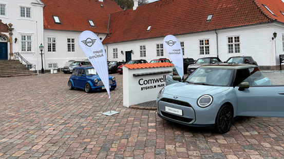 To MINI'er holder parkeret uden for en bygning med røde tagsten og brostensbelægning ved siden af et Comwell Bygholm Park-skilt.