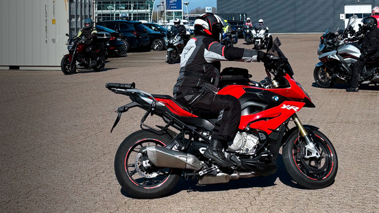 En person i fuldt køreudstyr sidder på en rød BMW XR-motorcykel på en parkeringsplads med andre motorcyklister i nærheden.