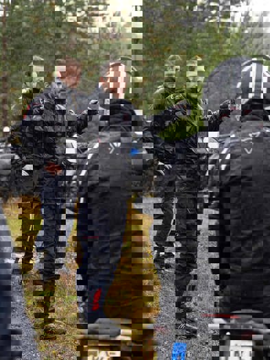 En gruppe af mennesker, med motorcykler iført motorcykeltøj og hjelme, står udenfor i en skov, imens en fortæller.