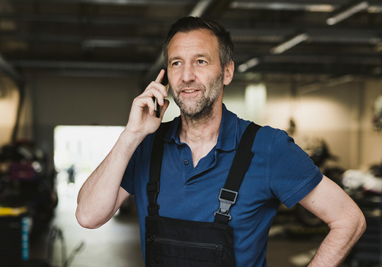 En mand i arbejdsdragt taler i mobiltelefon inde i en garage eller et værksted, hvor man kan se køretøjer og mennesker i baggrunden.