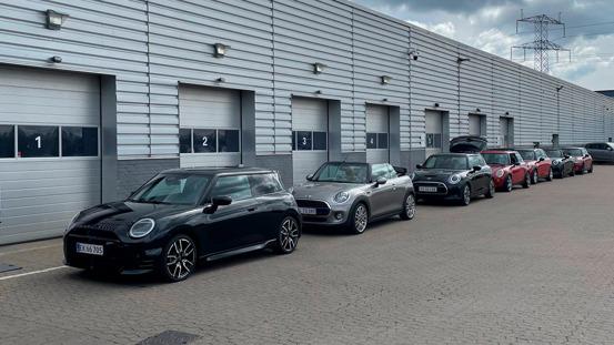 Seks Mini Cooper-biler af forskellige modeller står pænt parkeret på rad og række uden for en bygning med nummererede garageporte.