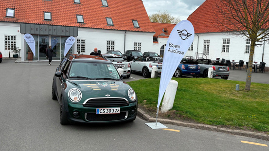 En grøn Mini Cooper parkeret ved et Bayern AutoGroup-flag ved Comwell Hotel Bygholm PArk i Horsens med andre biler og mennesker i baggrunden.