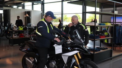 To mænd står ved siden af en BMW-motorcykel i et moderne showroom med tøj og hjelme pænt udstillet i baggrunden.