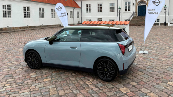 En lyseblå 3-dørs hatchback-bil parkeret på en brostensbelagt plads i nærheden af to Bayern AutoGroup-flag og en hvid bygning.