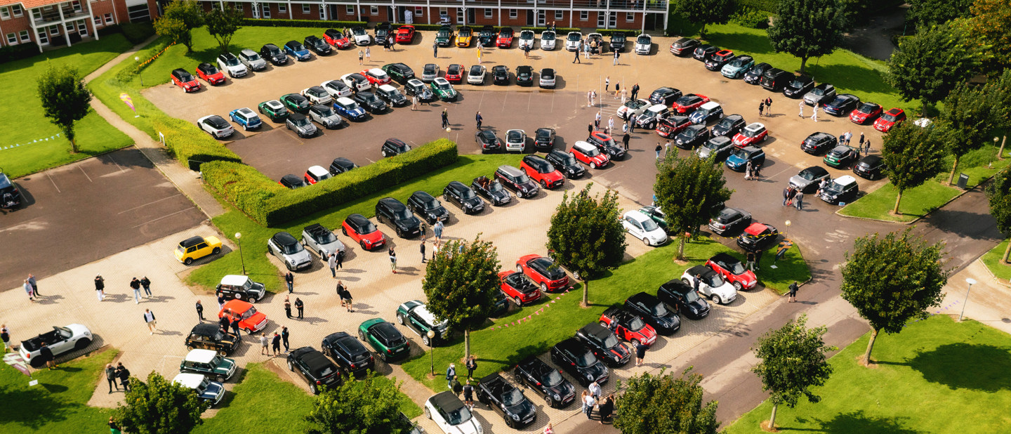 Luftfoto af en parkeringsplads fyldt med biler, med grupper af mennesker samlet mellem træer og græsarealer.