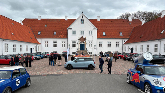 Folk samles omkring forskellige MINI-biler på gårdspladsen til en stor hvid historisk bygning med rødt tegltag.