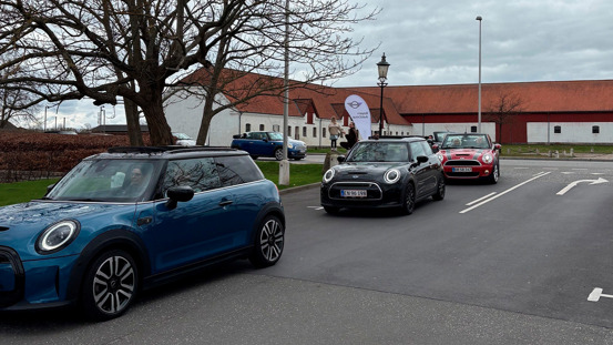 Tre Mini Coopers, blå, sort og rød, er parkeret på række på en asfalteret vej i nærheden af hvide bygninger og bladløse træer.