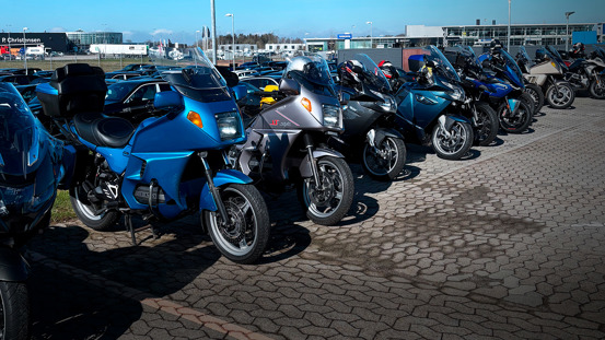 En række parkerede touring-motorcykler er opstillet på en asfalteret plads under en klar himmel med bygninger og biler i baggrunden.