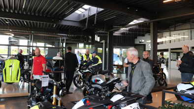 Flere mennesker diskuterer og ser på motorcykler i et moderne showroom med store vinduer og industri-inspireret indretning.
