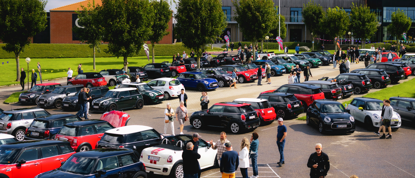 Flere mennesker er udendørs på en solrig dag, omgivet af parkerede Mini Coopers i forskellige farver.