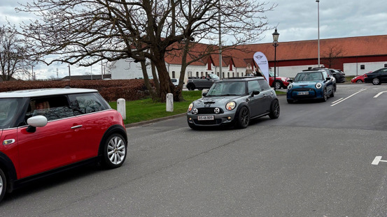 En række Mini Coopers kører på en gade i nærheden af en bygning med rødt tag og bladløse træer på en overskyet dag.