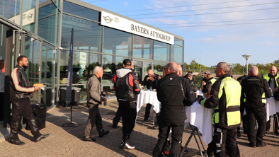 En gruppe mennesker i motorcykeludstyr samles udendørs nær indgangen til en bygning i Bayern AutoGroup, hvor de står ved høje borde.
