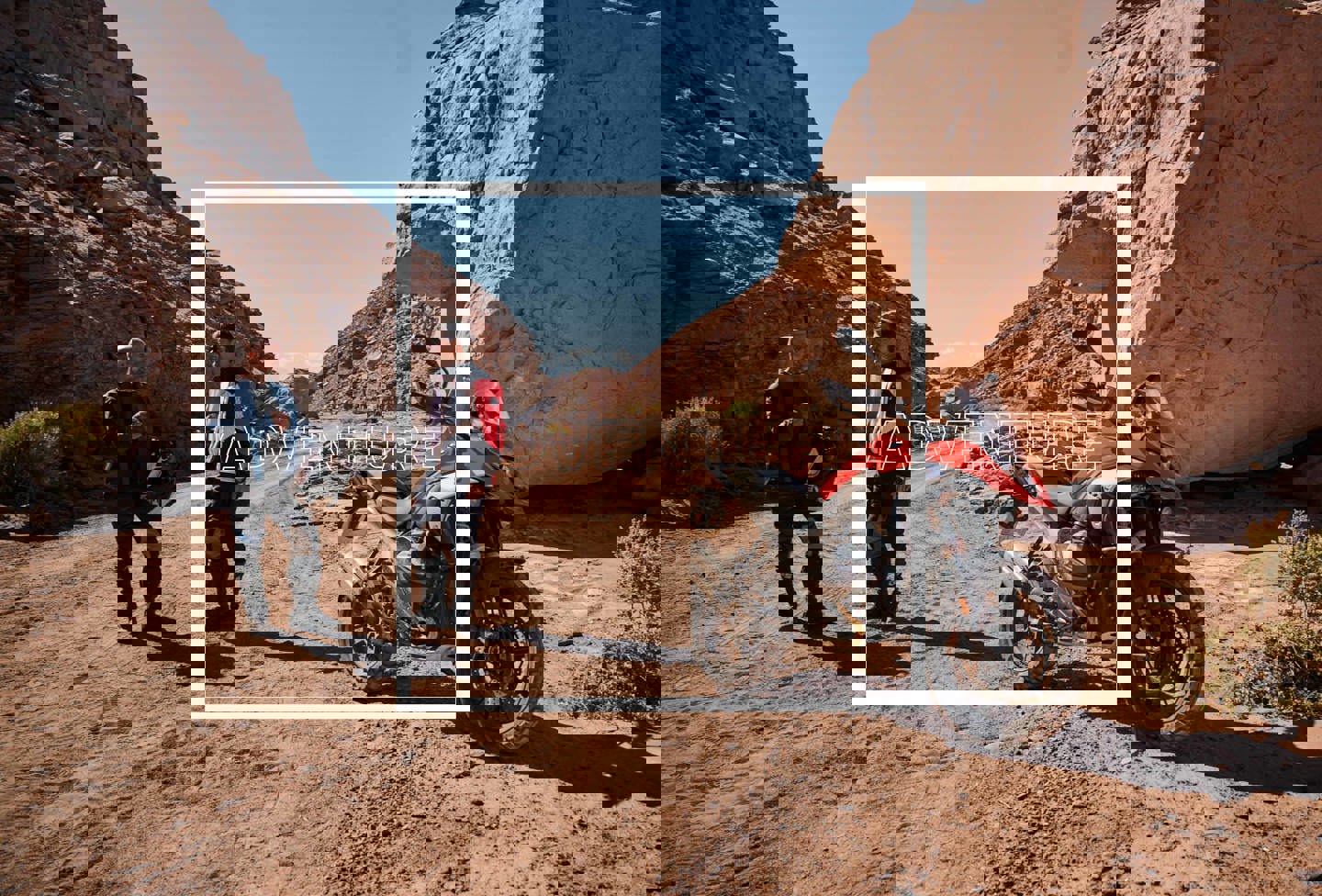 To personer står bagved en motorcykel i ørkenen. Henover billedet er der skrevet ADVENTURE med hvid.