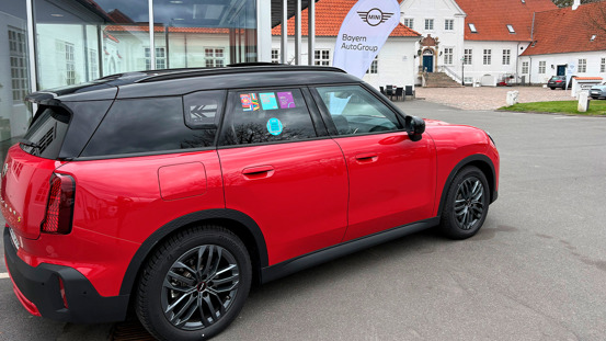 En rød og sort MINI-bil parkeret uden for en forhandlerbygning med et skilt, hvor der står Bayern AutoGroup MINI.