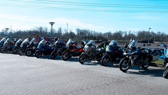 En række parkerede motorcykler pænt opstillet på et asfalteret område med træer og en åben mark i baggrunden.