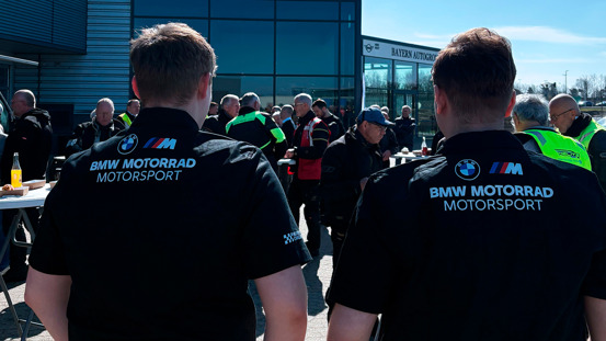 To personer iført BMW Motorrad Motorsport-trøjer står sammen med andre udenfor i nærheden af en moderne bygning.