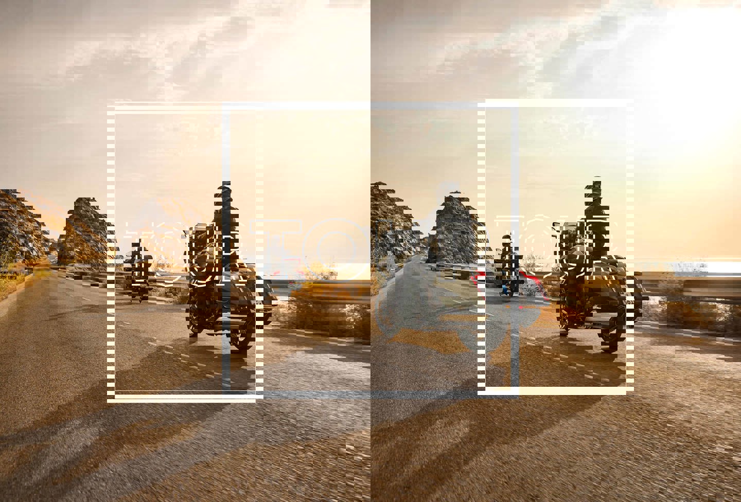 To personer kører på motorcykler på en vej med bjerge, vand og solskin i baggrunden. Der er en grafisk tekstboks på billede med TOUR skrevet i hvid. 