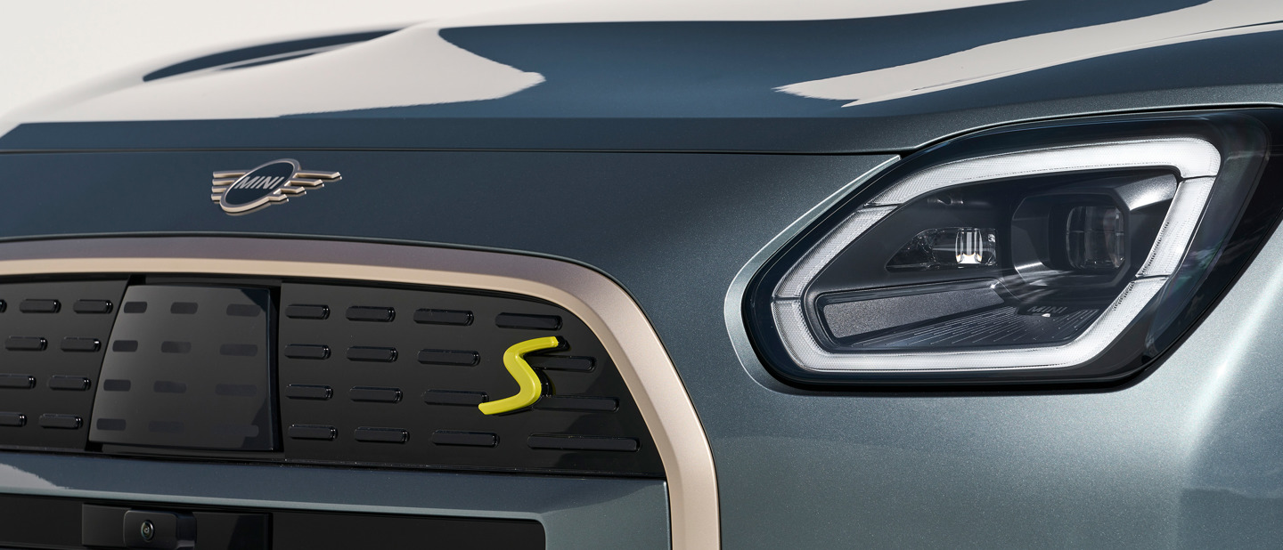 Nærbillede af fronten på en MINI-elbil, der viser forlygten, kølergrillen og MINI-logoet med et gult S-mærke.