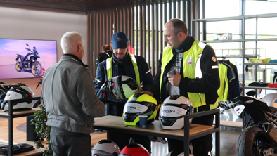Tre mænd iført motorcykeludstyr kigger på hjelme ved et udstillingsbord i et moderne motorcykeludstillingslokale.