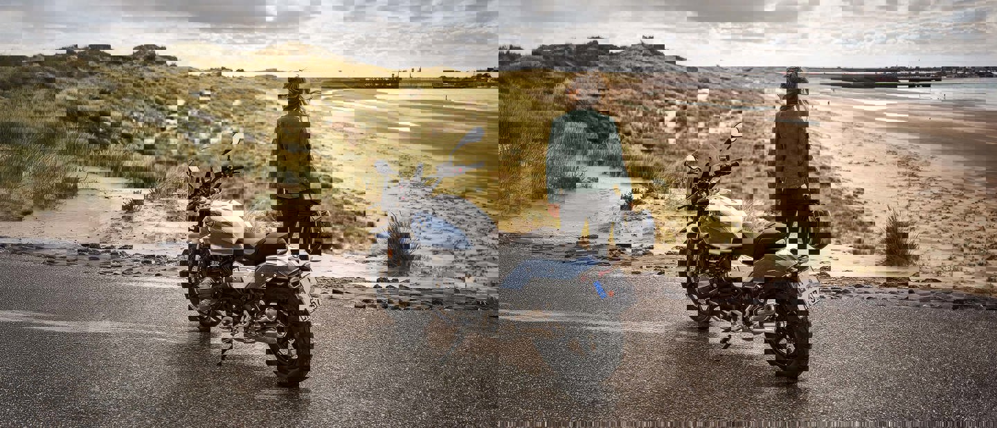 En person står ved siden af en motorcykel på en vej med sandklitter og en overskyet himmel i baggrunden.