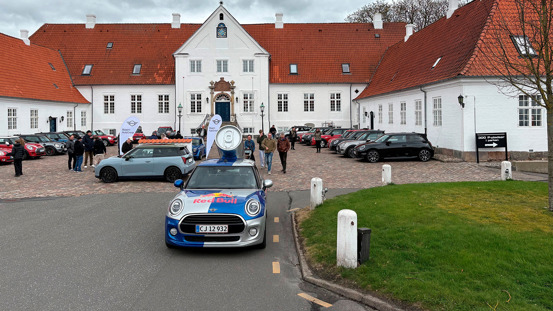 En Red Bull Mini-bil er parkeret foran en stor hvid bygning med rødt tag, omgivet af andre biler og mennesker.