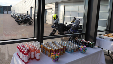 Et bord med diverse drikkevarer og snacks er arrangeret indendørs i nærheden af store vinduer med frit udsyn til parkerede motorcykler udenfor.