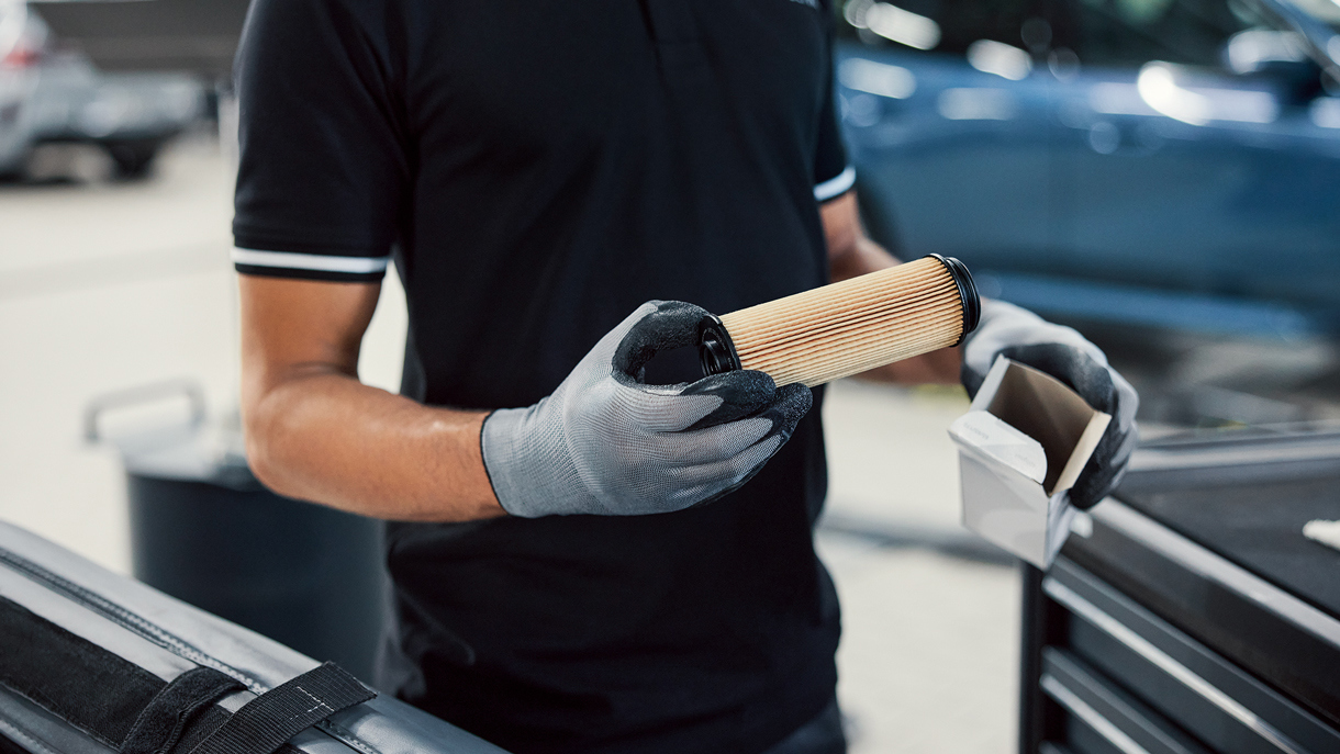 En person iført handsker holder et cylindrisk bilfilter ved siden af en værktøjskasse på et bilværksted.
