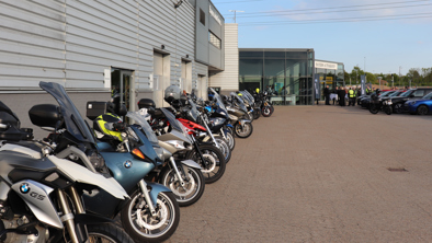 En række parkerede motorcykler uden for en moderne bygning med folk samlet ved indgangen.