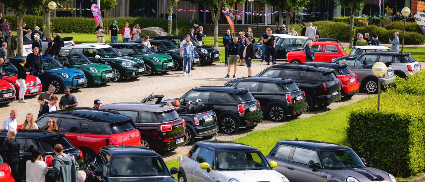 Folk samles på en parkeringsplads, hvor der vises forskellige modeller af Mini Cooper-biler, med grønne områder og bygninger i baggrunden.