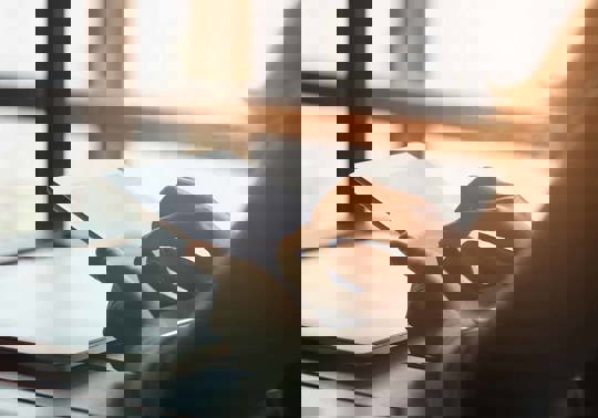 En person læser på sin ipad, indendørs i nærheden af store vinduer, hvor sollyset strømmer ind.