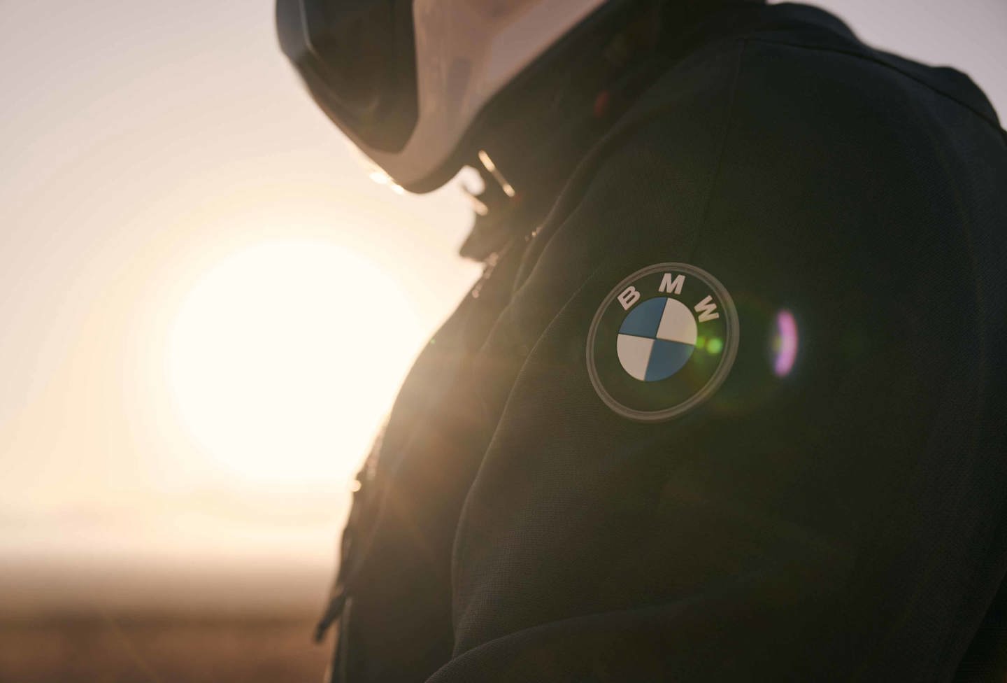 Et nærbillede af en person iført motorcykeljakke og hjelm med BMW Logo på armen, med solen i baggrunden. 