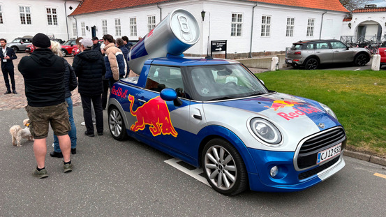 En Mini Cooper med Red Bull-branding og en stor dåse på taget er parkeret på en gade i nærheden af en gruppe mennesker.
