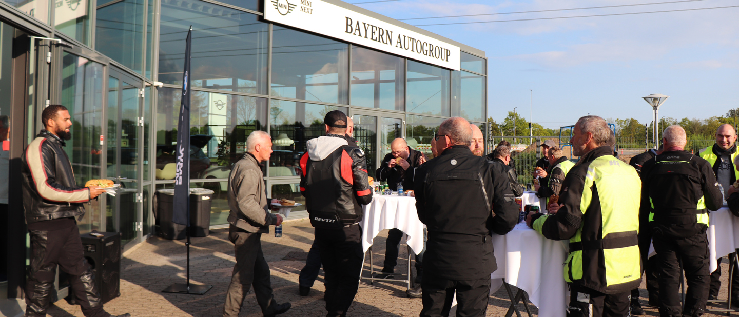 En gruppe motorcyklister samles uden for Bayern Autogroup, hvor de sludrer og står omkring bordene i deres køredragt.