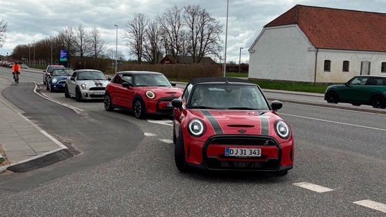 En række Mini Coopers kører ned ad en bygade med en cyklist på fortovet og træer i baggrunden.